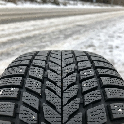 Close-up of a winter tire tread pattern, showing deep grooves and sipes for snow and ice traction, with a blurred snowy road background.
